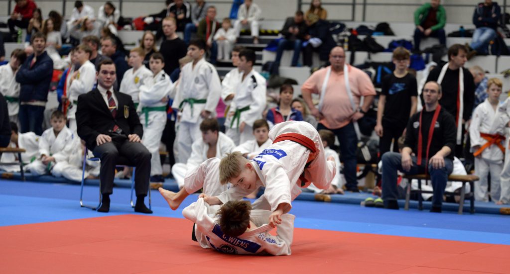 „Zwei junge Judoka im weißen Judogi kämpfen auf einer roten Matte beim 4. Hermann-Cup des PSV Schwerin, während ein Kampfrichter und Zuschauer am Mattenrand das Geschehen verfolgen.“