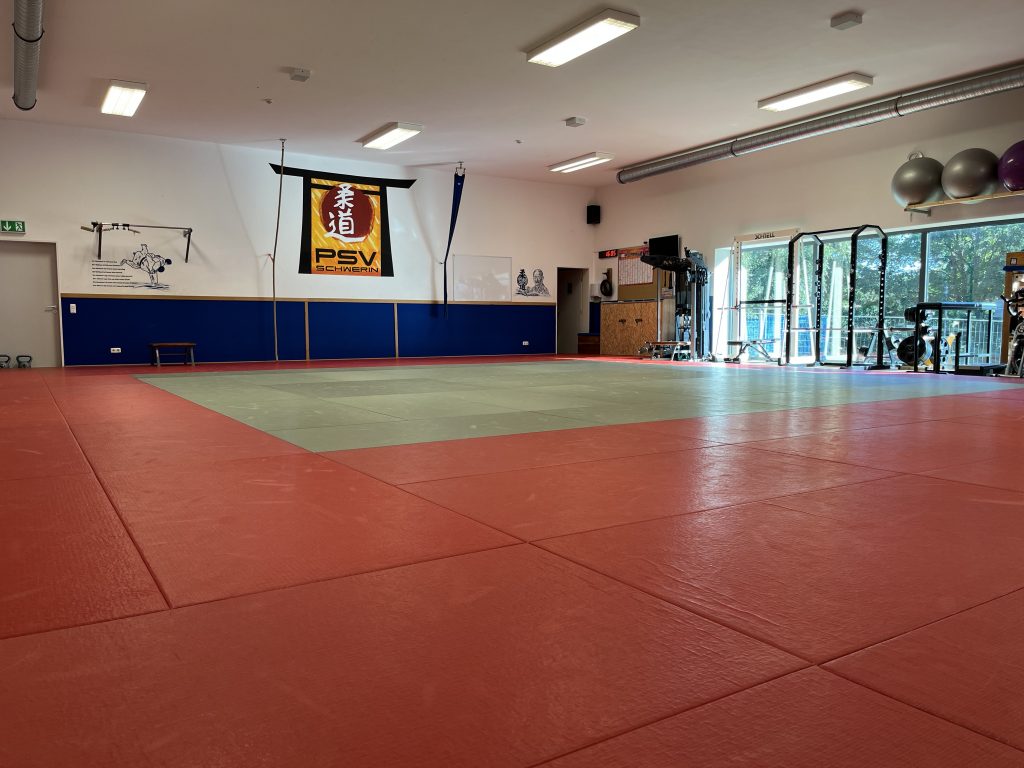Innenansicht eines Dojos mit roten und grünen Matten, Wandbanner „PSV Schwerin“ und Trainingsgeräten am Rand.
